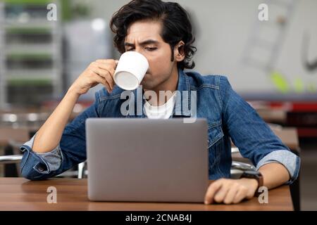 Portrait of a young handsome Indian man sipping from a coffee mug, sitting in an office cafeteria, coffee shop, casual work environment. Stock Photo