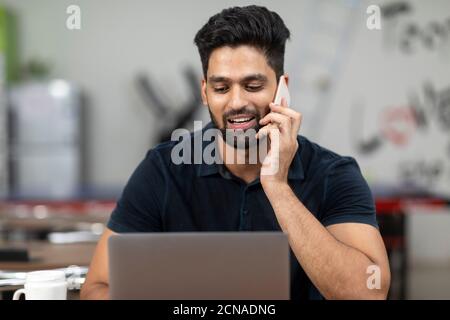 Portrait of a young handsome Indian man speaking on his phone and working on his laptop, sitting in an office cafeteria, coffee shop, casual work. Stock Photo