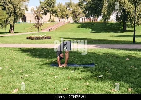 Young athletic man in sportswear doing yoga in the park. Practice standing forward bend asana pose outdoors. People exercising on green grass with Stock Photo