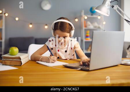 Online school training. Little girl in headphones is studying a lesson lecture having a laptop video call with teacher sitting at a table at home. Stock Photo