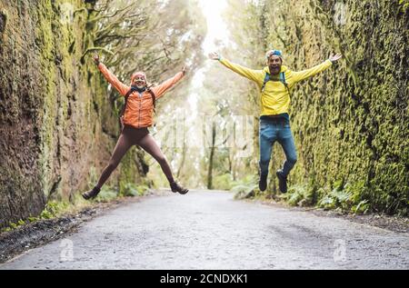 Happy couple jumping in touristic park - Young tourist having fun doing excursion in wild forest Stock Photo