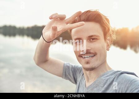 portrait of Young handsome stylish smiling guy makes selfie against the lake. peace sign Stock Photo