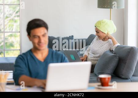 Asian businesswoman with towel binding on head sitting and using notebook computer on sofa, blur Caucasian businessman using laptop  working at desk. Stock Photo