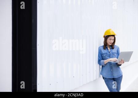 Beautiful Asian female engineer in jeans dress wearing yellow safety hard hat using laptop computer notebook doing job at construction site outside of Stock Photo
