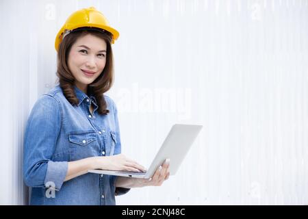 Beautiful Asian female engineer in jeans dress wearing yellow safety hard hat using laptop computer notebook doing job at construction site outside of Stock Photo