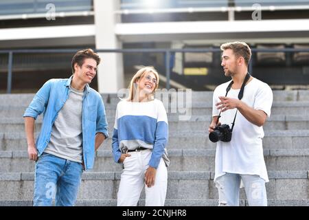 Three happy friends talking taking a conversation on the street in a sunny day with buildings in the background. Stock Photo