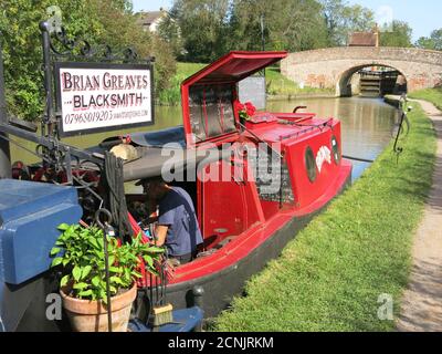 Traditional blacksmith, Brian Greaves, works on board his narrowboat on the Grand Union canal and sells his metalwork from the towpath near Braunston. Stock Photo