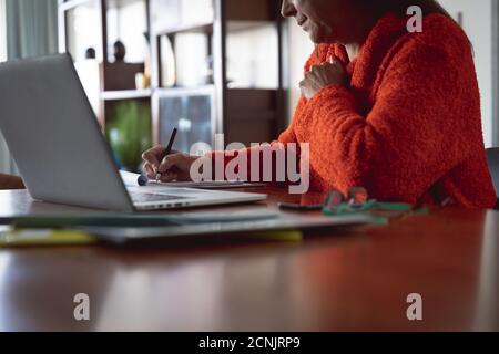 Caucasian woman enjoying time at home, social distancing and self isolation in quarantine lockdown, sitting by table, using a laptop and making notes. Stock Photo