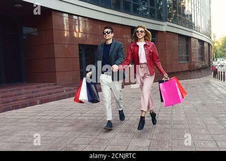 Front view of a casual couple of shoppers running in the street towards camera holding colorful shopping bags. Stock Photo