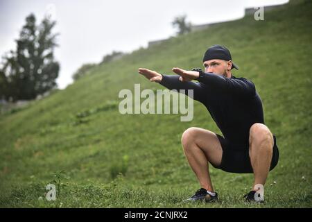 Young caucasian man performing squats before jogging on footpath in park. Stock Photo