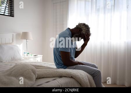 African American senior man sitting on a bed in a bedroom, holding his forehead in his hand, social distancing and self isolation in quarantine lockdo Stock Photo