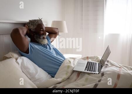 African American senior man lying on a bed in a bedroom, using a laptop and smiling, social distancing and self isolation in quarantine lockdown Stock Photo