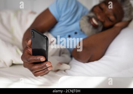 African American senior man lying on a bed in a bedroom, using a smartphone and smiling, social distancing and self isolation in quarantine lockdown Stock Photo
