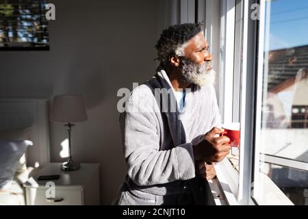 African American senior man standing in a bedroom, holding a cup of coffee, looking through a window, social distancing and self isolation in quaranti Stock Photo