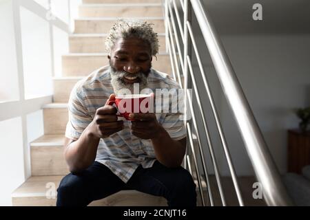 African American senior man sitting on stairs, holding a cup of coffee and smiling, social distancing and self isolation in quarantine lockdown Stock Photo