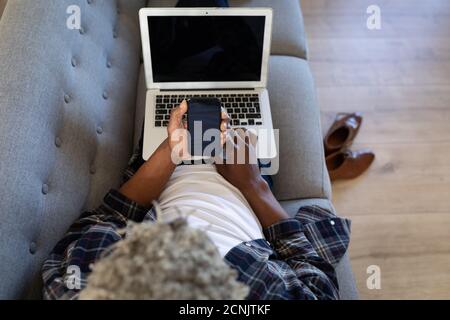 African American senior man lying on a couch, using a laptop, talking on a phone, social distancing and self isolation in quarantine lockdown Stock Photo