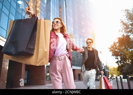 Front view of a casual couple of shoppers running in the street towards camera holding colorful shopping bags. Stock Photo