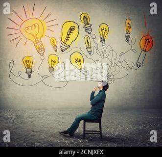 Businessman sitting on chair, keeps hand under chin, thoughtful gesture, looking up with interest, thinking of new ideas as multiple light bulbs come Stock Photo