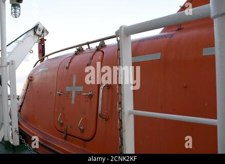 Close view on the orange polyurethane color life boat with reflective tape stickers secured with davits and safety hooks on the merchant cargo vessel. Stock Photo