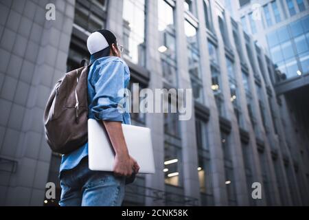 Cheerful young man with backpack enjoying walk the city with laptop and snap back.