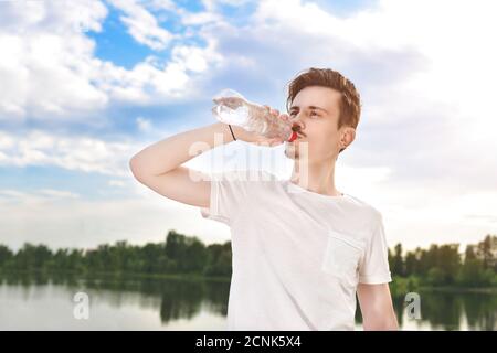 young guy drinks fresh water against of the lake and the forest background. summer thirst. Guy wants to drink Stock Photo