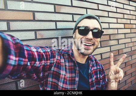 Cheerful stylish man in sunglasses and gray hat takes a selfie, standing against the brown brick wall. Stock Photo