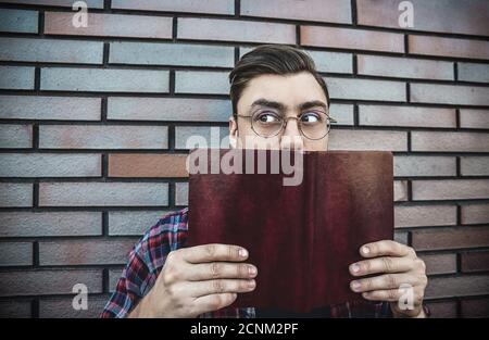 Young hipster guy wearing eyeglasses reading a book or note book on brick wall background. Stock Photo