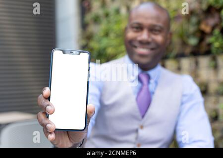 Executive black man showing a smartphone with a blank screen. He is wearing a medical mask and formal wear. Selective focus. Space for text. Business Stock Photo