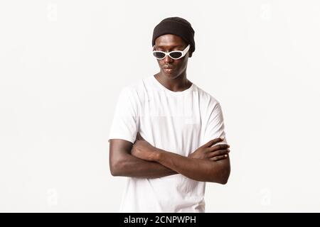 Portrait of handsome and stylish, sassy african-american guy in sunglasses and beanie, standing with arms crossed and posing over white background Stock Photo