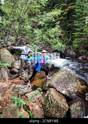 Asian friends hiking along Lost Lake Trail, having a good time embracing nature near a running stream. Popular place near Boulder, Colorado Stock Photo