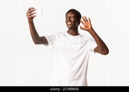 Portrait of happy smiling african-american guy talking with friends via video call, holding smartphone and waving hand in hello gesture, standing Stock Photo