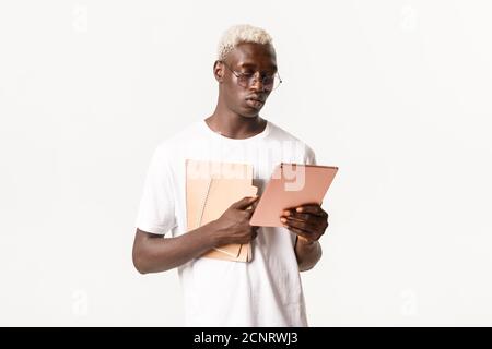 Portrait of determined, serious-looking african american male student, looking focused at digital tablet, studying, holding notebooks, standing white Stock Photo