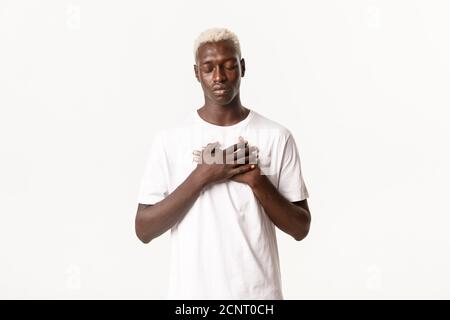 Portrait of nostalgic calm african-american blond man, holding hands on heart with closed eyes, standing white background Stock Photo