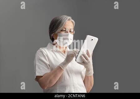 Doctor in protective mask work digital gadget. Adult Caucasian woman seriously looks at screen of white tablet. Cut out on gray background Stock Photo
