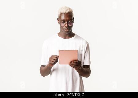 Portrait of handsome and stylish serious african-american man in glasses, looking at digital tablet, using device focused, standing white background Stock Photo