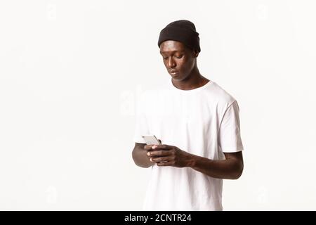 Portrait of serious attractive and cool african-american guy in beanie, messaging, looking at mobile phone, standing white background Stock Photo