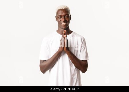 Portrait of smiling african-american handsome blond man, holding hands clasped together and looking thankful, standing white background Stock Photo