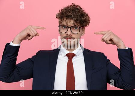 young elegant fashion man in navy blue suit wearing glasses and pointing fingers to head, standing on pink background Stock Photo