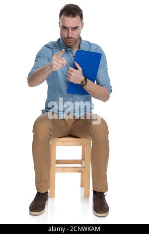 Furious smart casual man holding clipboard, pointing and frowning while sitting on a chair on white studio background Stock Photo
