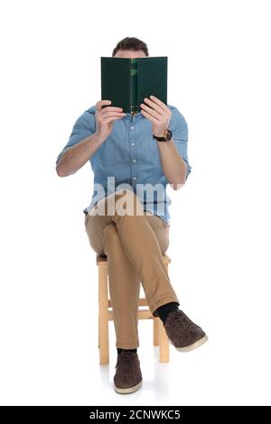 Young smart casual man covering his face with a book while sitting on a chair on white studio background Stock Photo