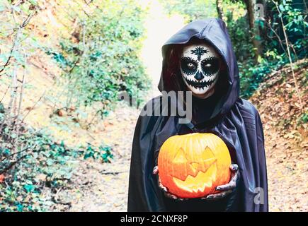 Young woman with halloween paint face mask wearing black hood - Scary witch holding spookey craved pumpkin Stock Photo