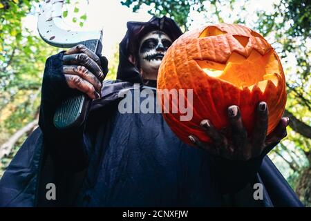 Young woman with halloween paint face mask wearing black hood - Scary witch holding spookey craved pumpkin Stock Photo