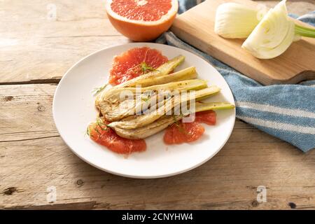 Sautéed fennel vegetable with grapefruit slices, vegetarian meal on a white plate, ingredients and a blue kitchen towel on rustic wooden planks, copy Stock Photo