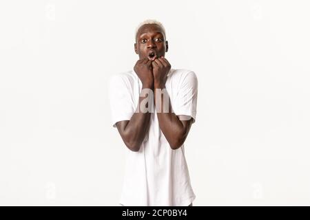 Portrait of amazed and fascinated african-american blond man, gasping excited, holding hands pressed to face, looking at camera thrilled, standing Stock Photo