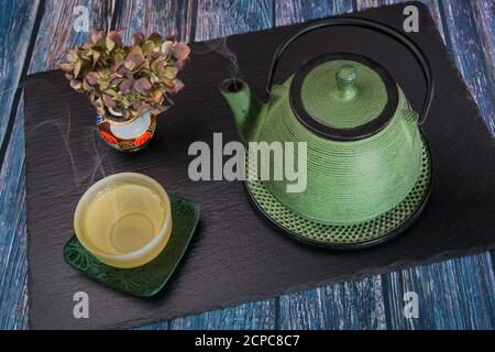 A steaming green metal teapot, a glass bowl with steaming tea and a chinese vase with dried hydrangea on a slate slab. Stock Photo