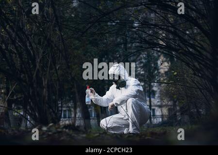 Laboratory assistant working in residential area, researching plastic litter, wearing protective coverall, gas mask and gloves, crouched in bushes Stock Photo