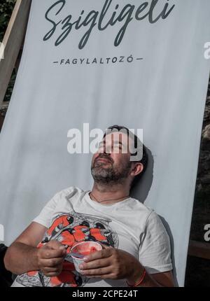 SZIGLIGET, HUNGARY - 08/26/2020: Caucasian man with closed eyes taking rest with ice cream. Sunny day. Stock Photo