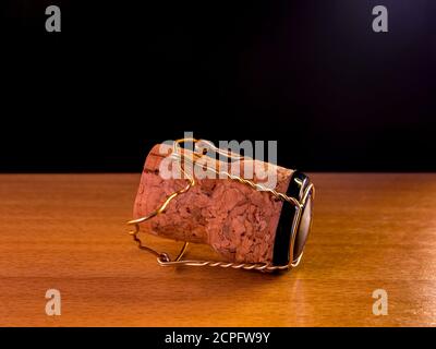 Cork plug with muselet(wire cage) from champagne or sparkling wine lies on a wooden surface with black background and a hint of lighting from the Stock Photo