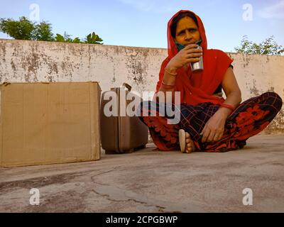 DISTRICT KATNI, INDIA - MAY 21, 2020: An indian village lady drinking water sitting with luggage at sky wall background. Stock Photo