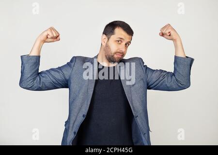 Young smiling businessman man with beard in jacket showing arm muscles, smiling proudly. Fitness concept. Portrait of a man on a gray background. Stock Photo